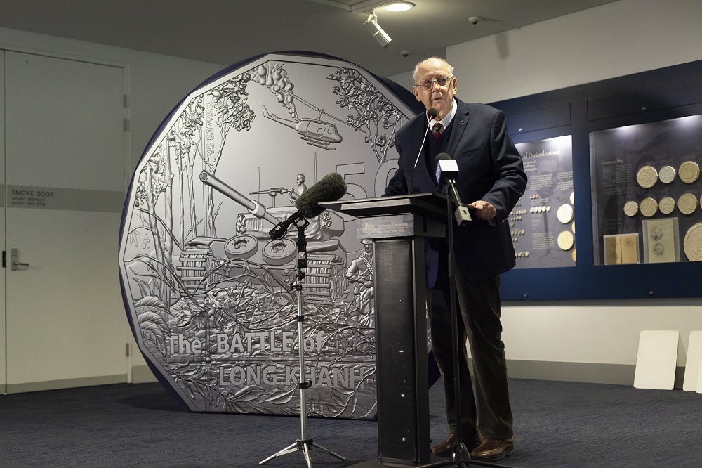 Elderly man at lectern with very large mock 50 cent coin behind him