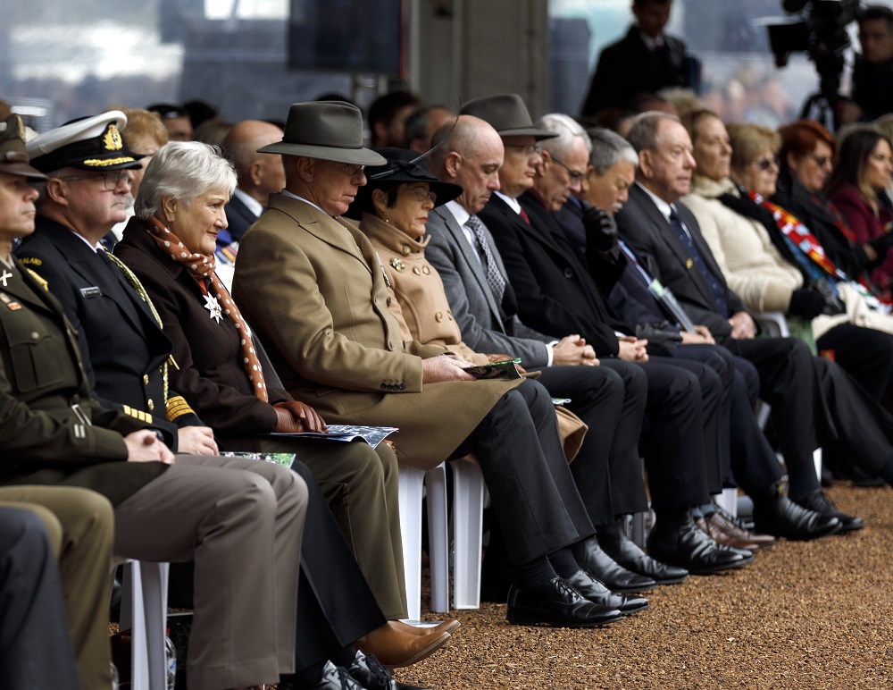 A row of people in suits and uniforms seated with heads bowed.