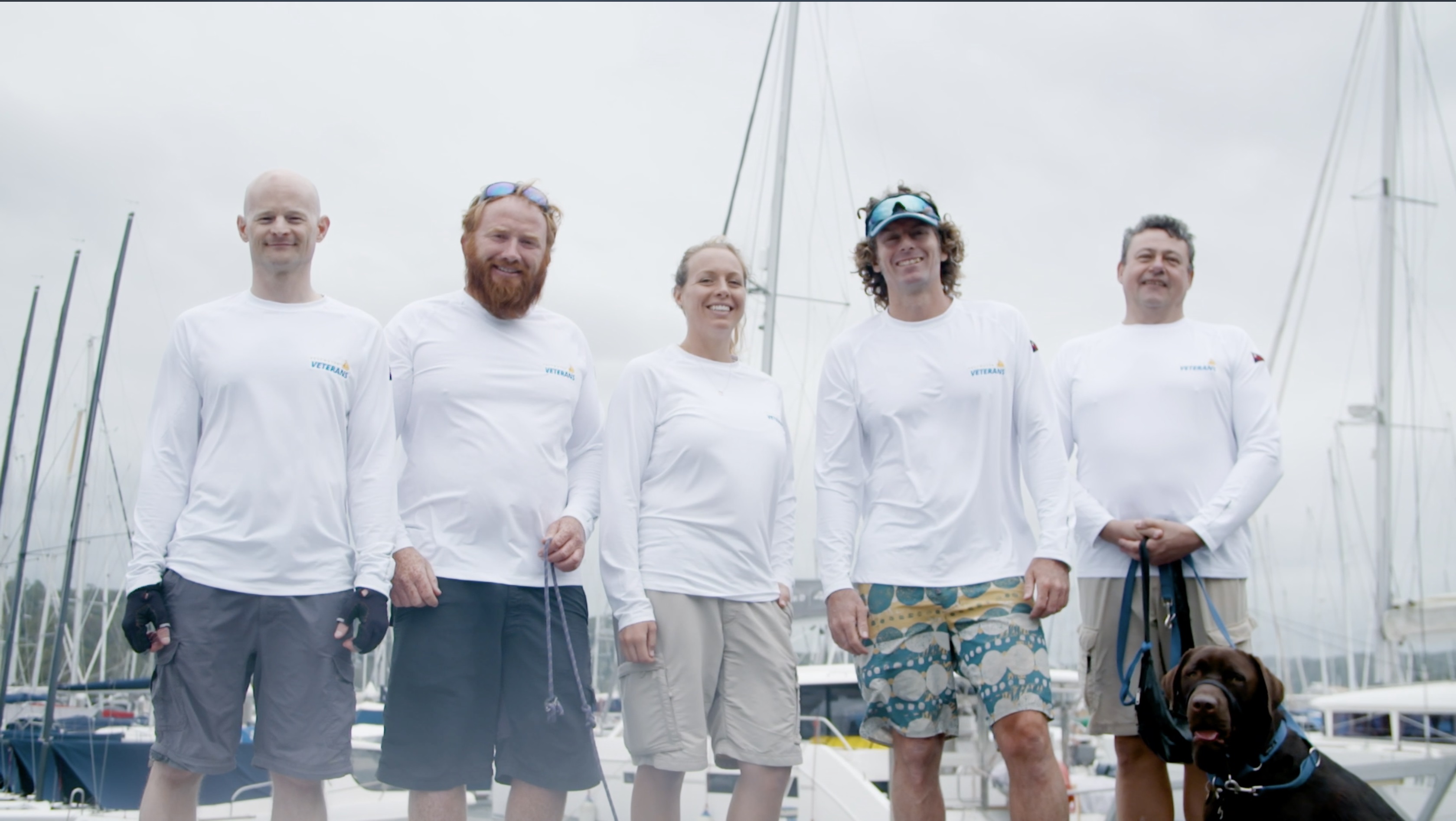 Four men, a woman and a dog posing with yachts in background.