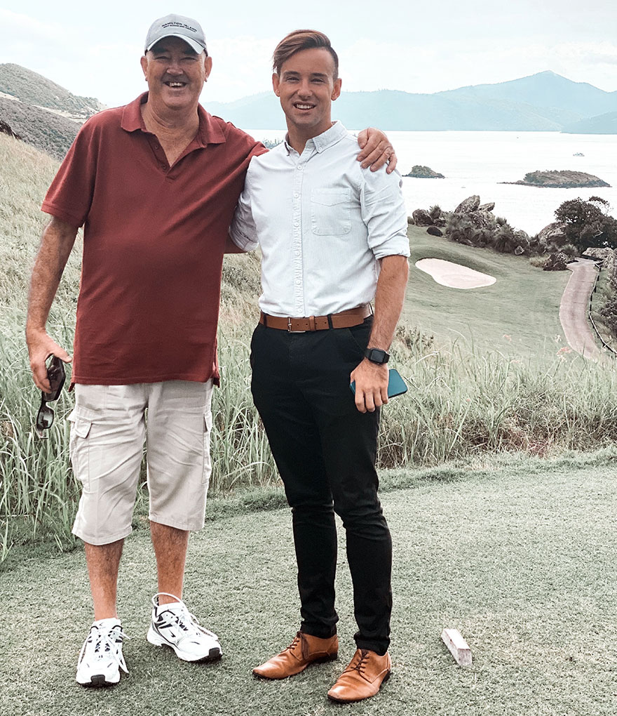 Dean Marchini standing with his dad. There is a sloping mountainside and sea in the background.