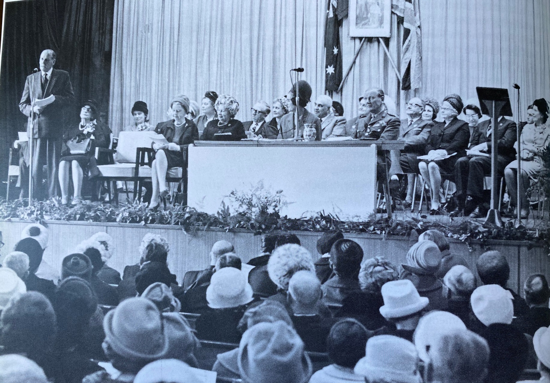 Black and white photo showing man on stage speaking to large group of women