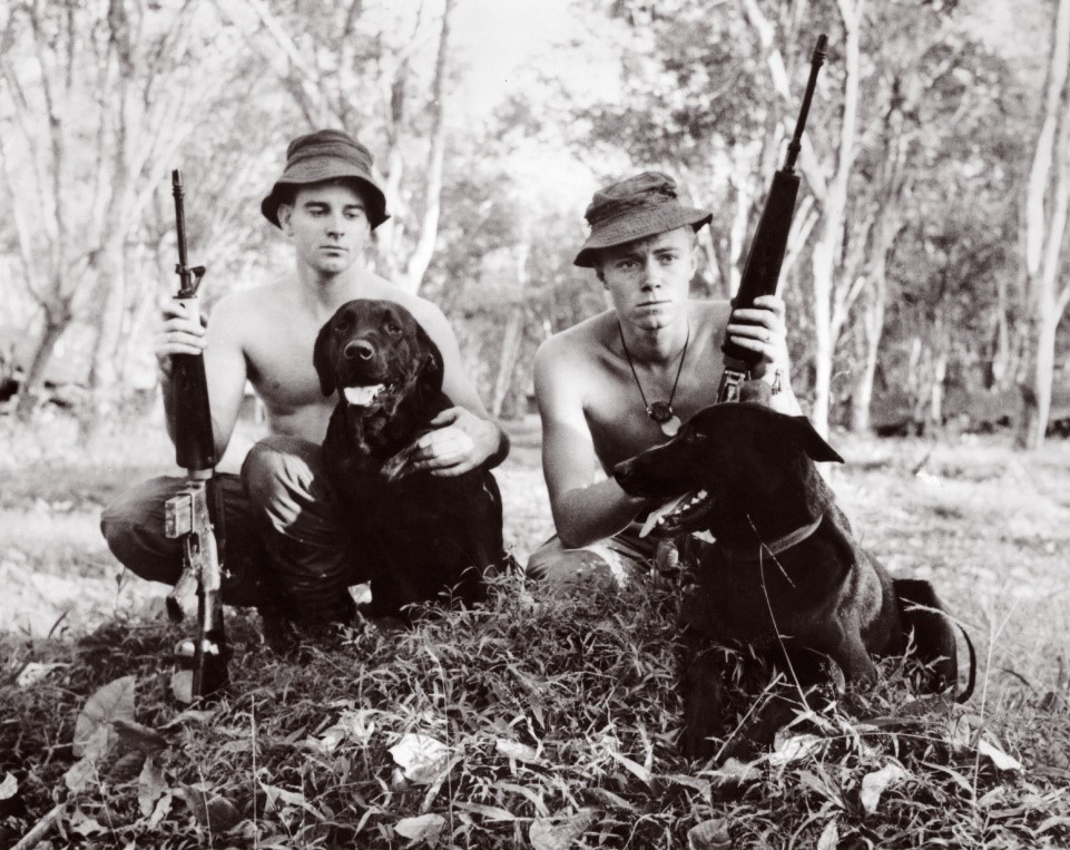 Two young men holding assault rifles with black labradors