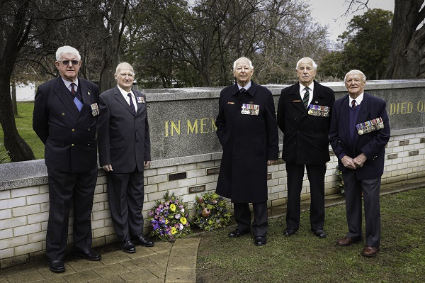 Five elderly men wearing medals stand in front of low wall with brass plaques attached