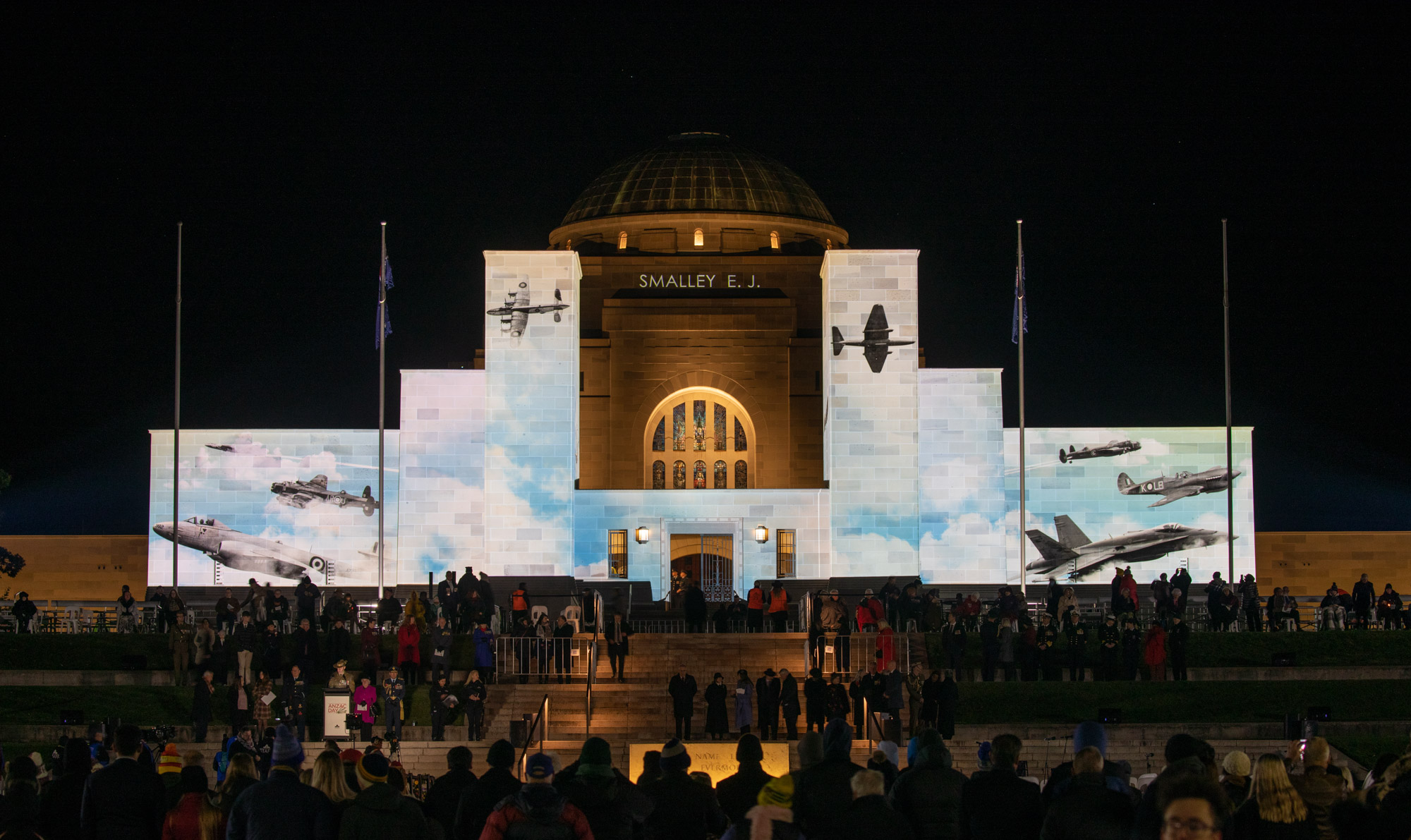 Night shot of front of AWM with crowd in foreground and various historic planes projected on the facade.