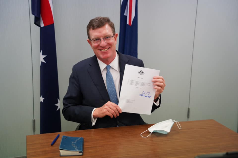 Middle-aged man in suit sitting a desk, smiling and holding document up to the camera.