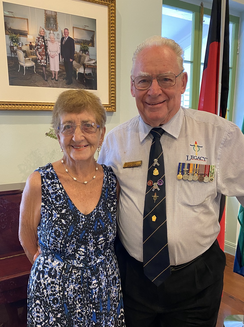 Elderly man and woman standing in front of a framed photo of the Queen