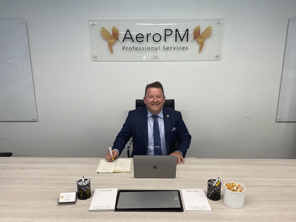 Smiling middle-aged man in office with sign reading AeroPM