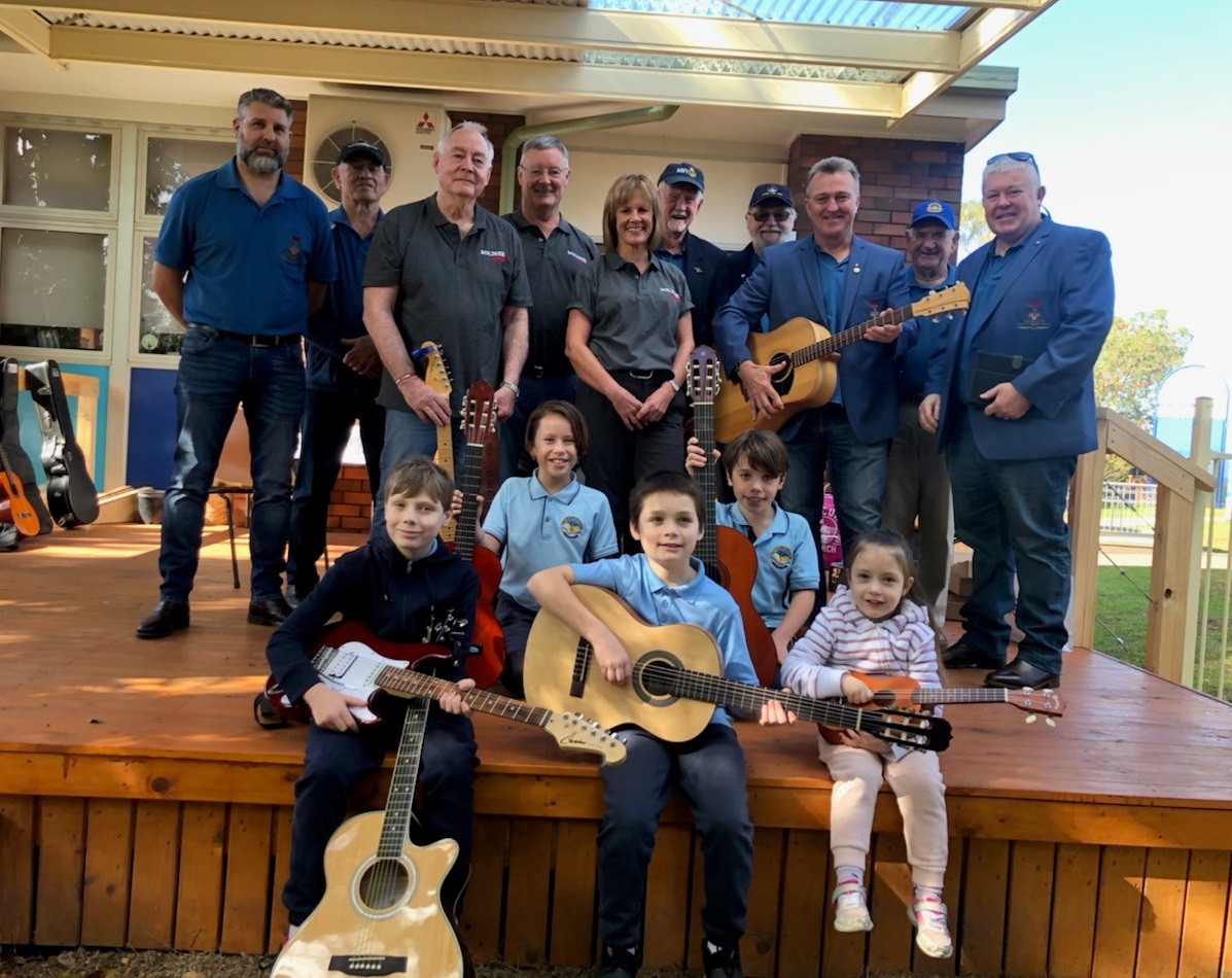 Several middle-aged people standing on an outside deck, with five children at their feet clutching guitars.