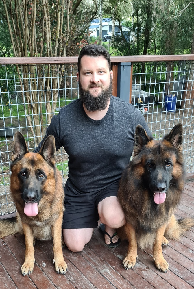 Bearded man crouching on timber deck between two German shepherds