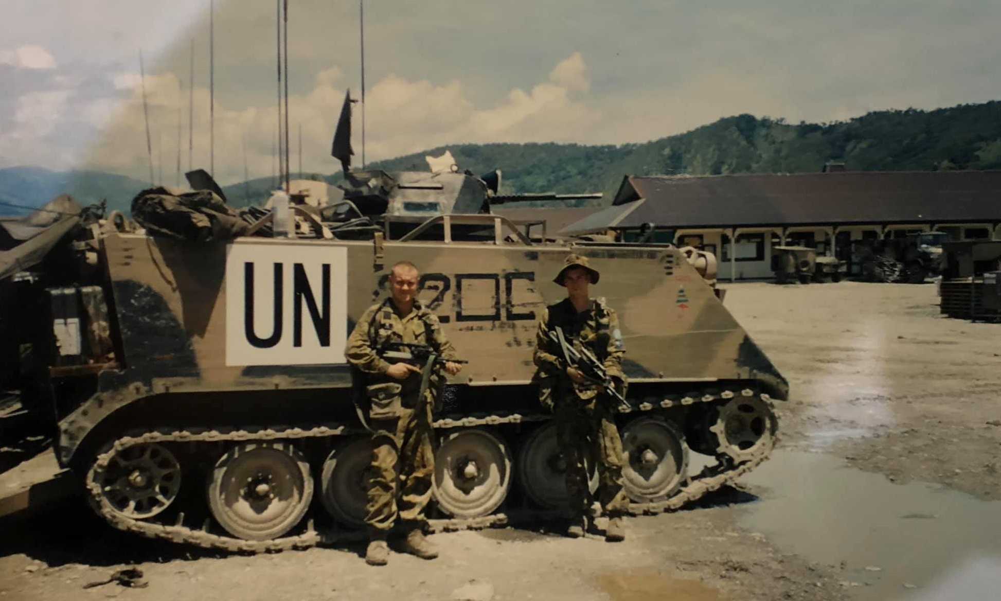 Two soldiers standing in front of armoured personnel carrier with hills in background