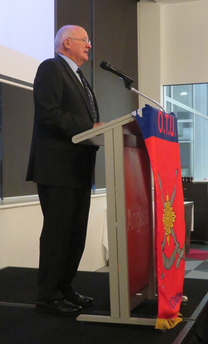 Ageing man standing at lectern with OTU banner hanging from its front