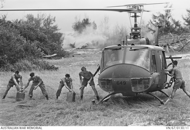 Five young soldiers unloading gerry cans from a Huey helicopter on an expanse of grass with bush in the background