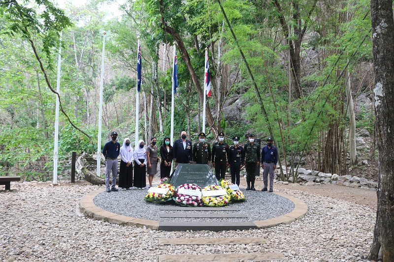 Long shot of about a dozen people in masks next large plaque with wreaths of flowers, flags and jungle in background