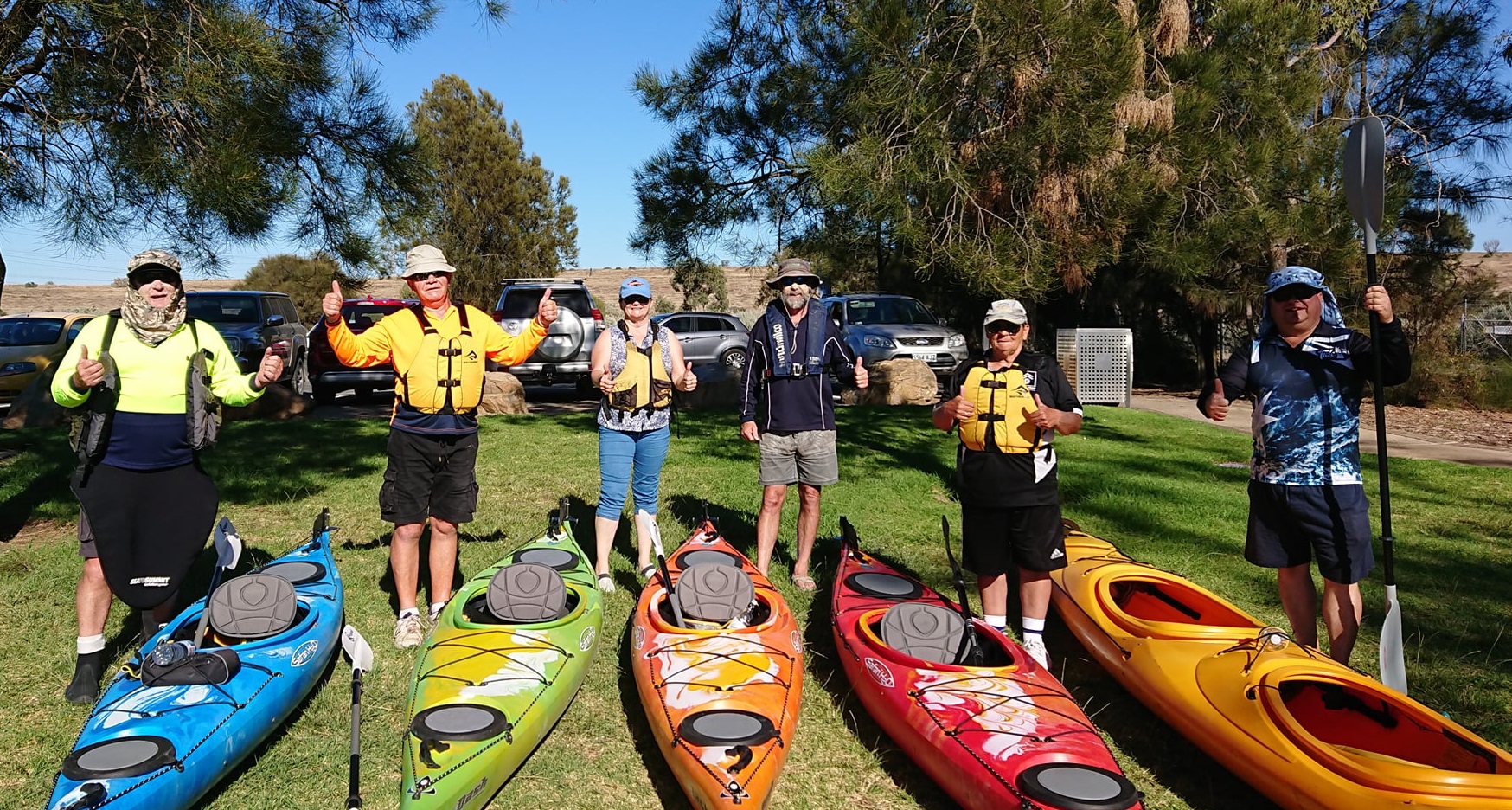 Six people pose next to their kayaks with thumbs up