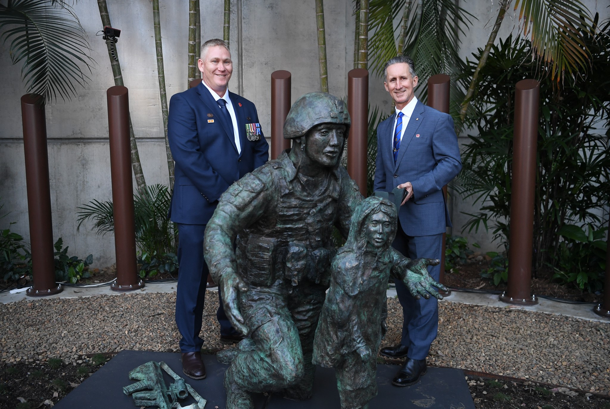 Two middle-aged men smile at camera with statue of soldier and girl in the foreground