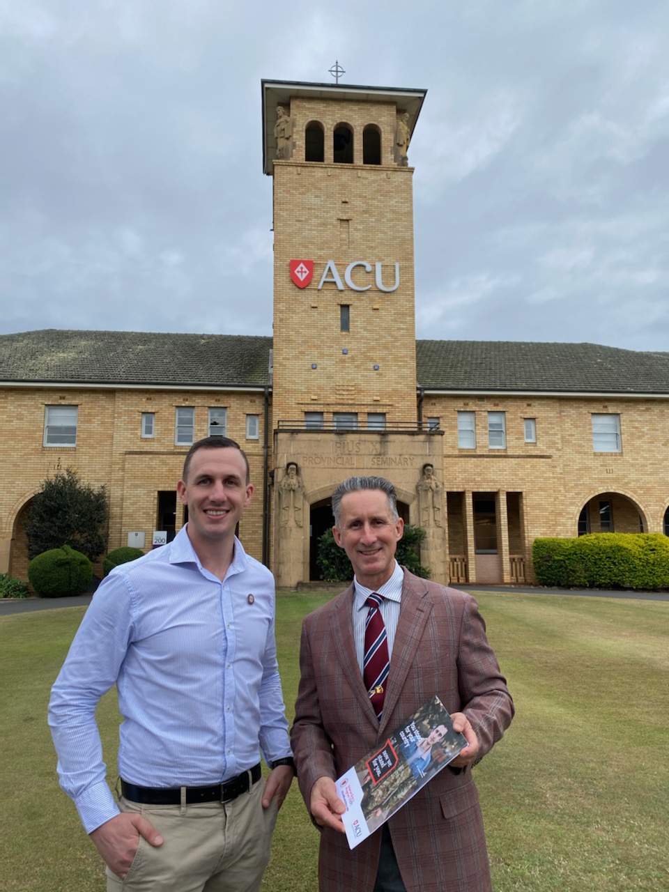 Two men posing for camera with building in background with letters ACU on it