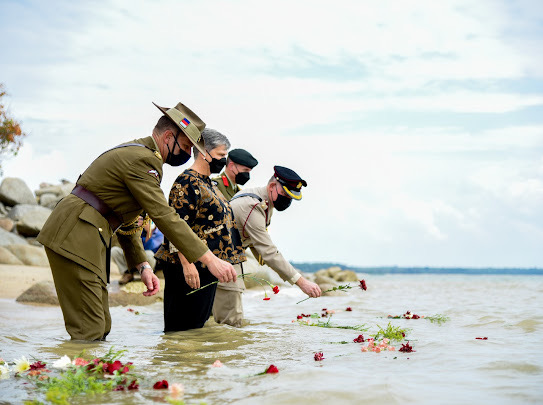 A civilian woman and three men in uniform knee-deep in the sea lay wreaths