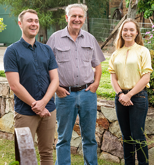 An older man with a young man on his right, and young woman on his left, all smiling, in a garden