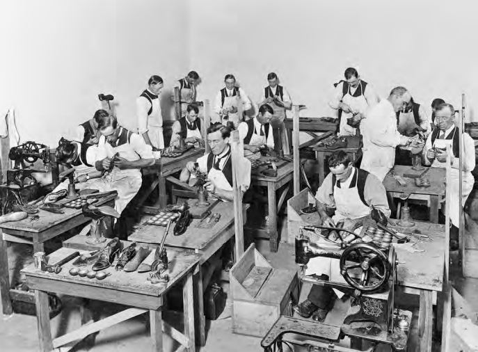 Black and white photo of a dozen men working at tables with boot-making equipment