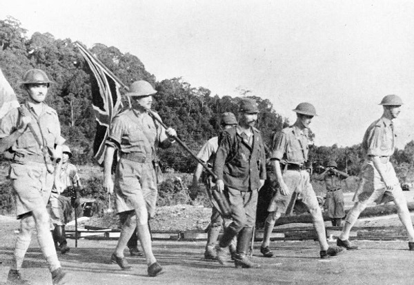 Four British soldiers, once carrying the Union Jack, march accompanied by Japanese officer