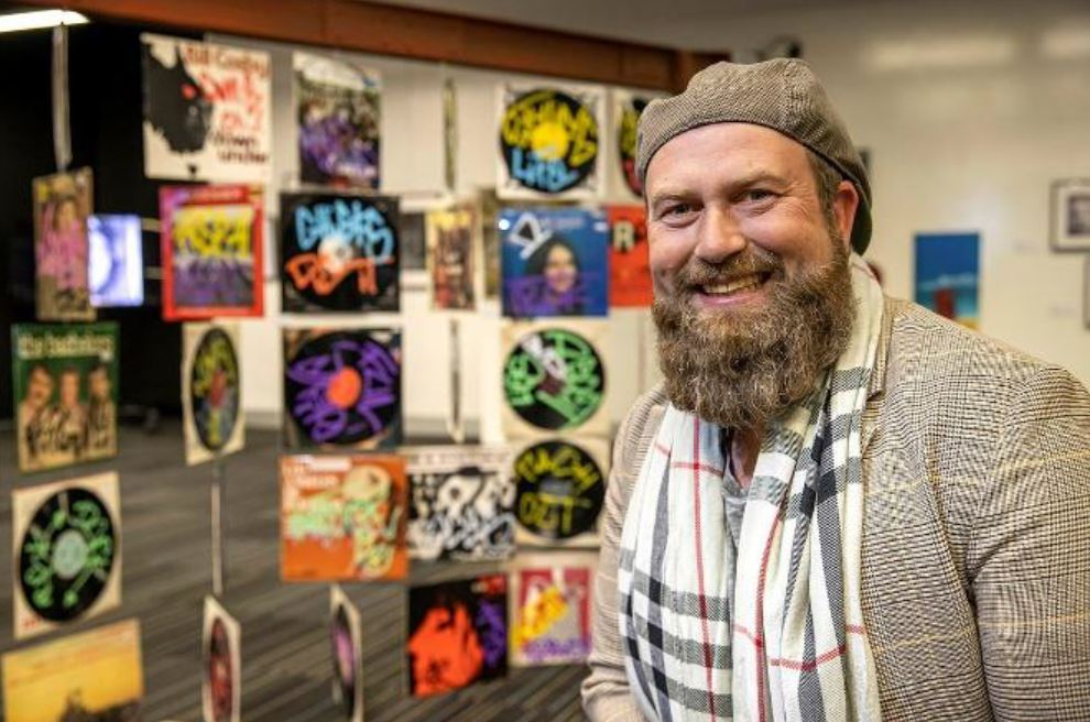 Bearded man in beret smiling next to art works hanging on wall.