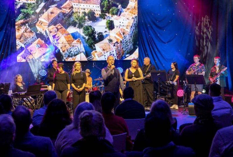 Several women on stage singing to audience, with musicians behind them.