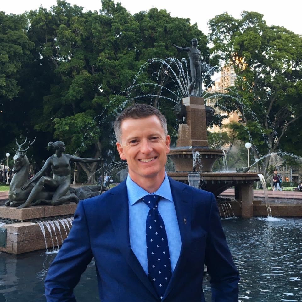 Man in suit smiling at camera with Sydney's Hyde Park fountain in background