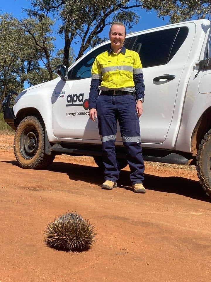 Woman on a red dirt road, standing in front of a Ute and smiling with an echidna in the foreground 