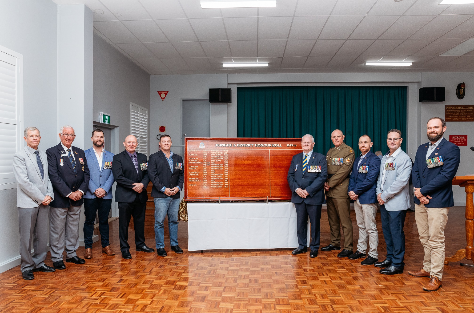 Ten men pose next to honour roll inside hall