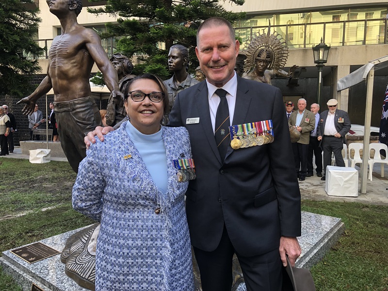 Middle-aged man and Indigenous woman, both wearing medals, in front of bronze sculpture