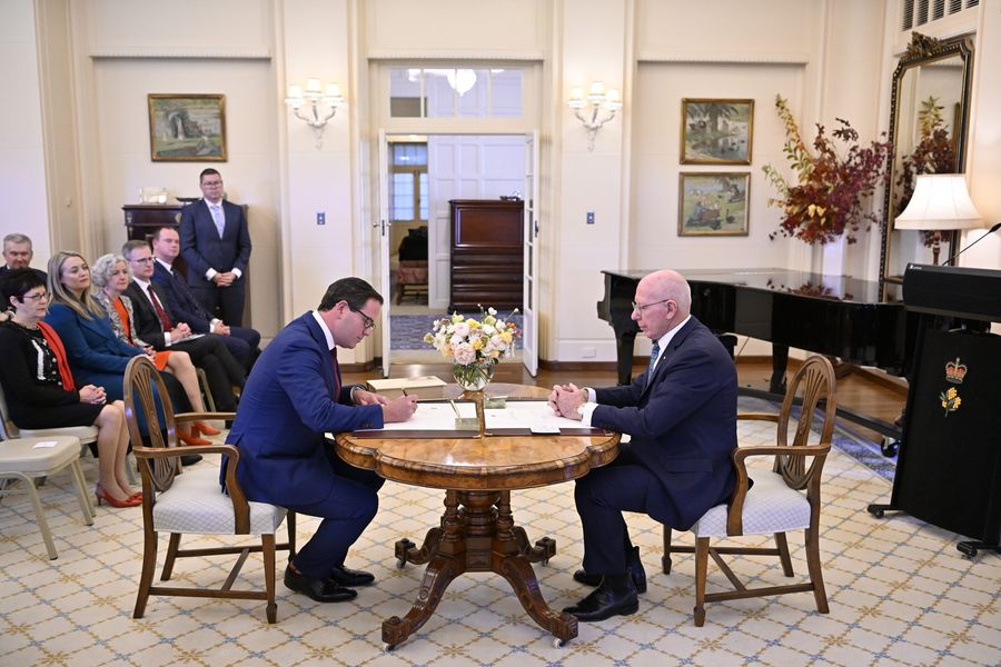 Man in suit writes on document on table with older man looking on and small audience watching