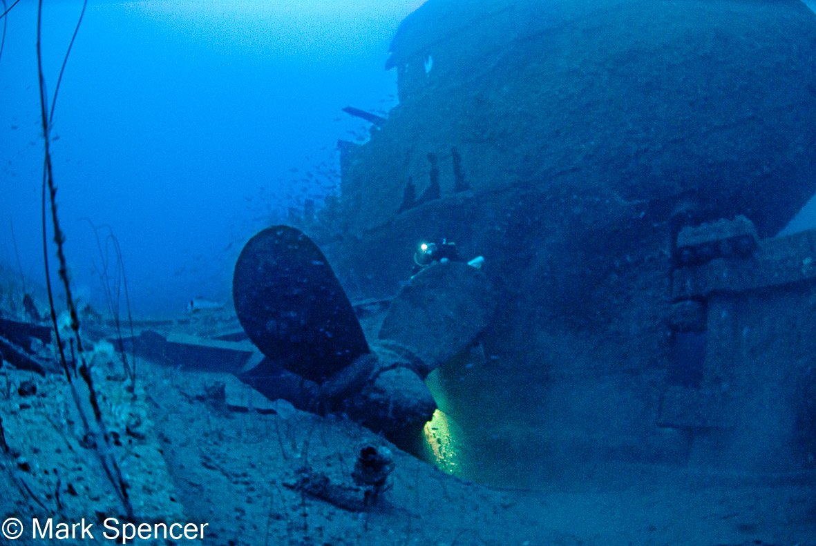 The stern of a sunken ship sits on the ocean floor. A large propeller sits in the foreground.