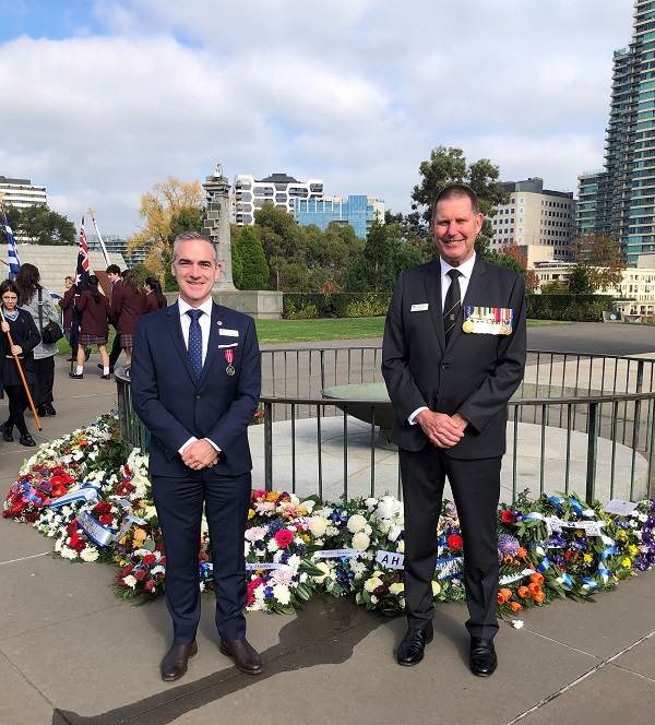 Two middle-aged men smile at camera in front of small memorial surrounded by wreaths