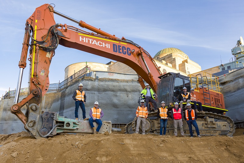 Eight men posing for camera on giant digger with Australian War Memorial in background