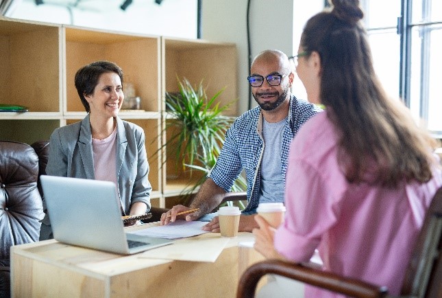 three people sitting at a office table with open laptop and one person holding coffee cup