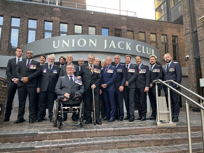 About 15 men in suits, one in a wheelchair, posing out the front of the Union Jack Club