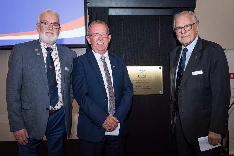 Three older men in suits pose smiling next to plaque