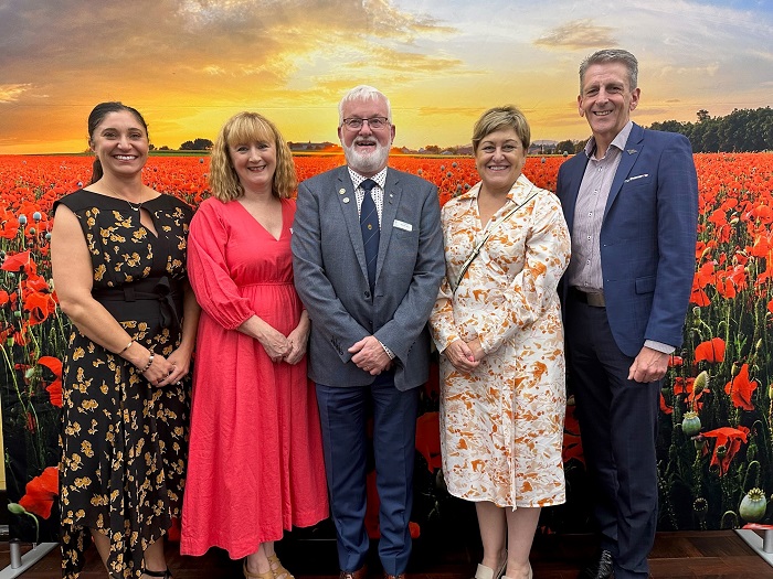 Three women and two men posing and smiling in front of wall photo of poppies