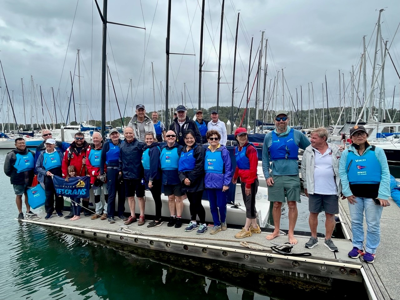 About 20 people stand on marina next to sailing boats