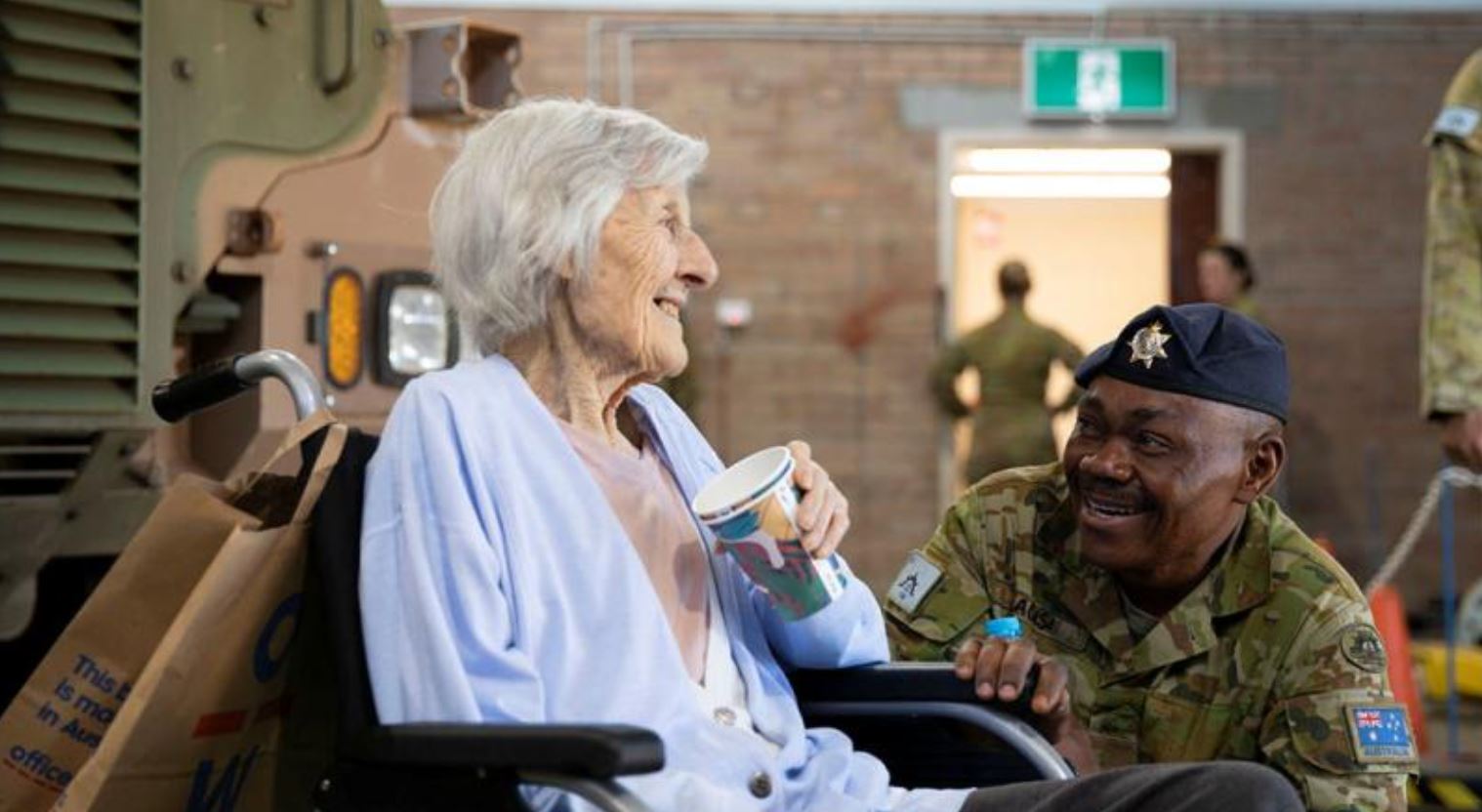 An elderly lady in wheelchair next to Army truck chats with soldier