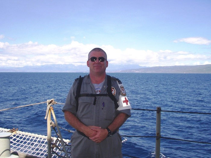 A man with sunglasses and dressed in grey overalls with a red cross symbol on a ship with the water and land in the distance