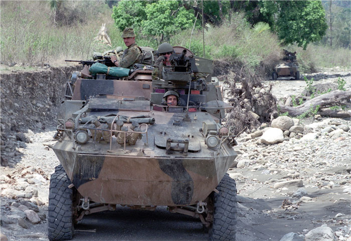 A tank with three soldiers, two are manning guns, travelling on a rubble road