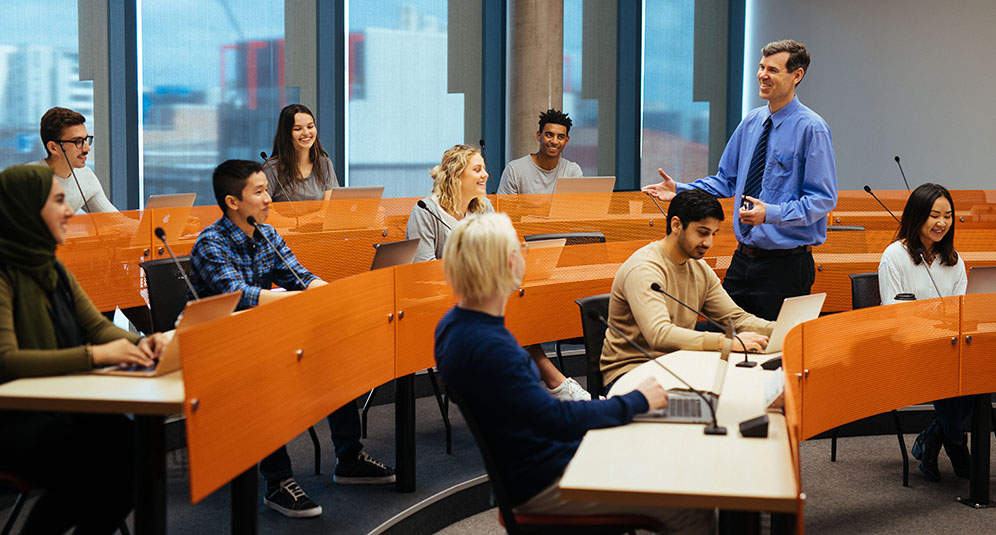 9 students of different ages and backgrounds sit at desks in a University of Newcastle room, listening to a lecturer.