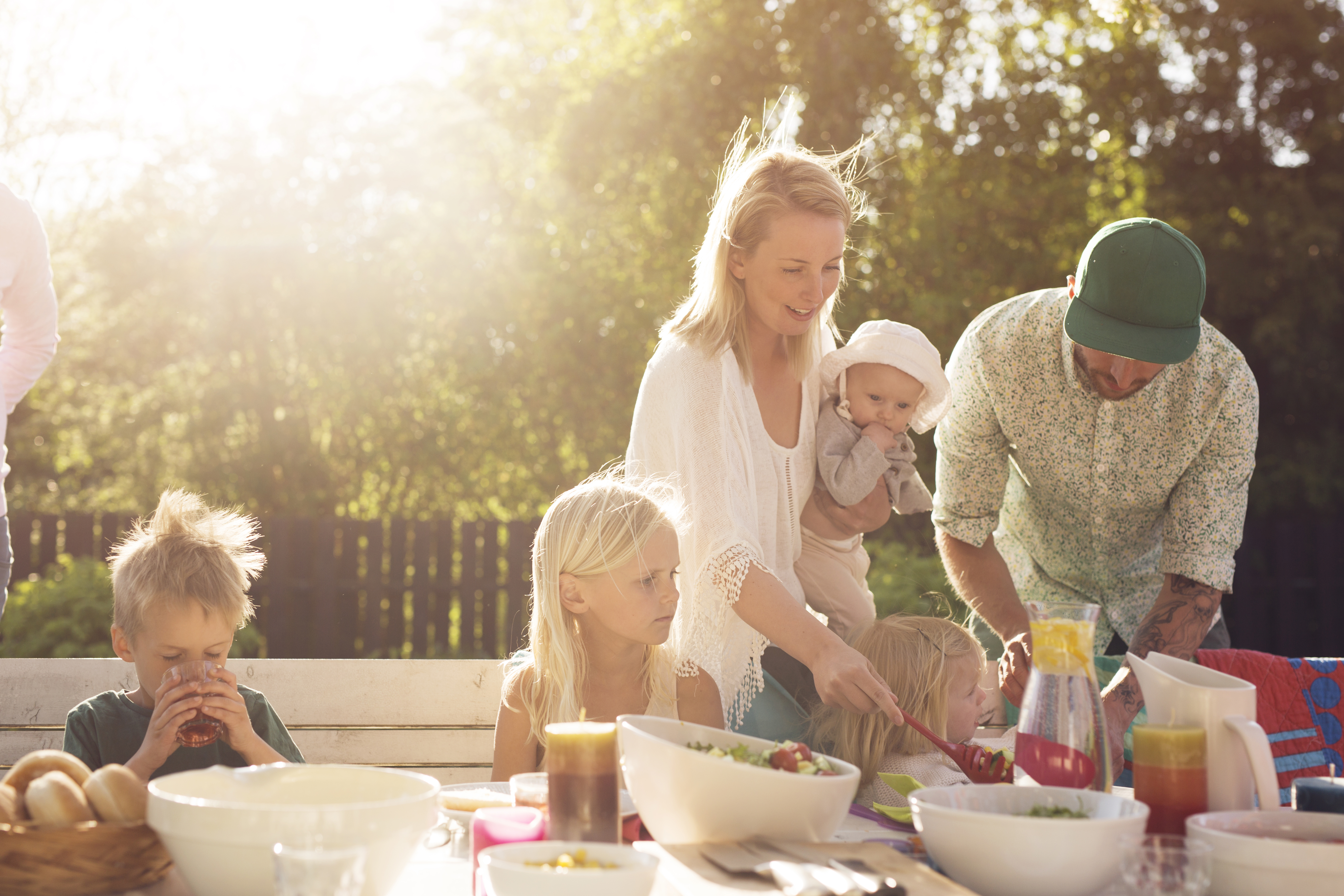 A family with a mother father and 3 young children are around an outdoor table having a picnic bbq style dinner as the sun is setting