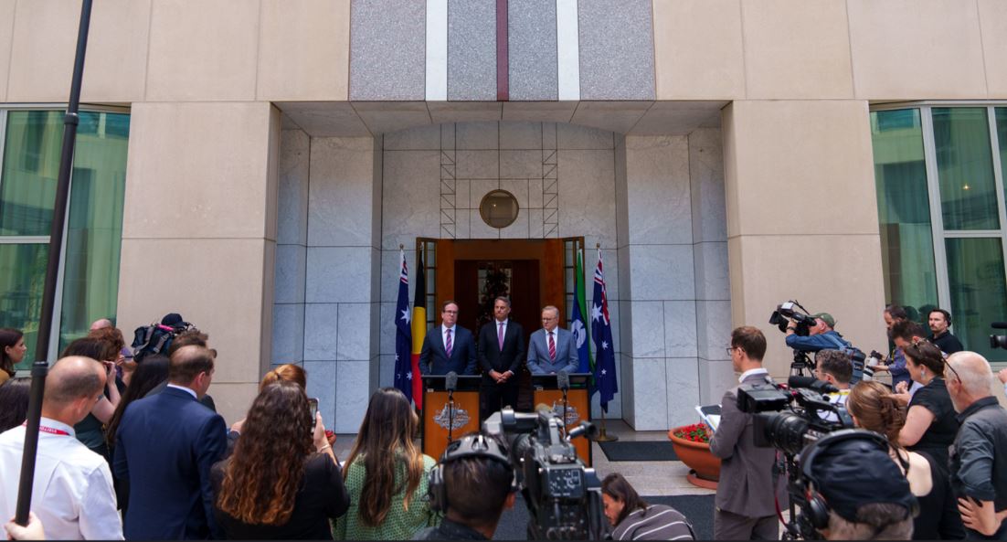 Three men in suits before gaggle of journalists