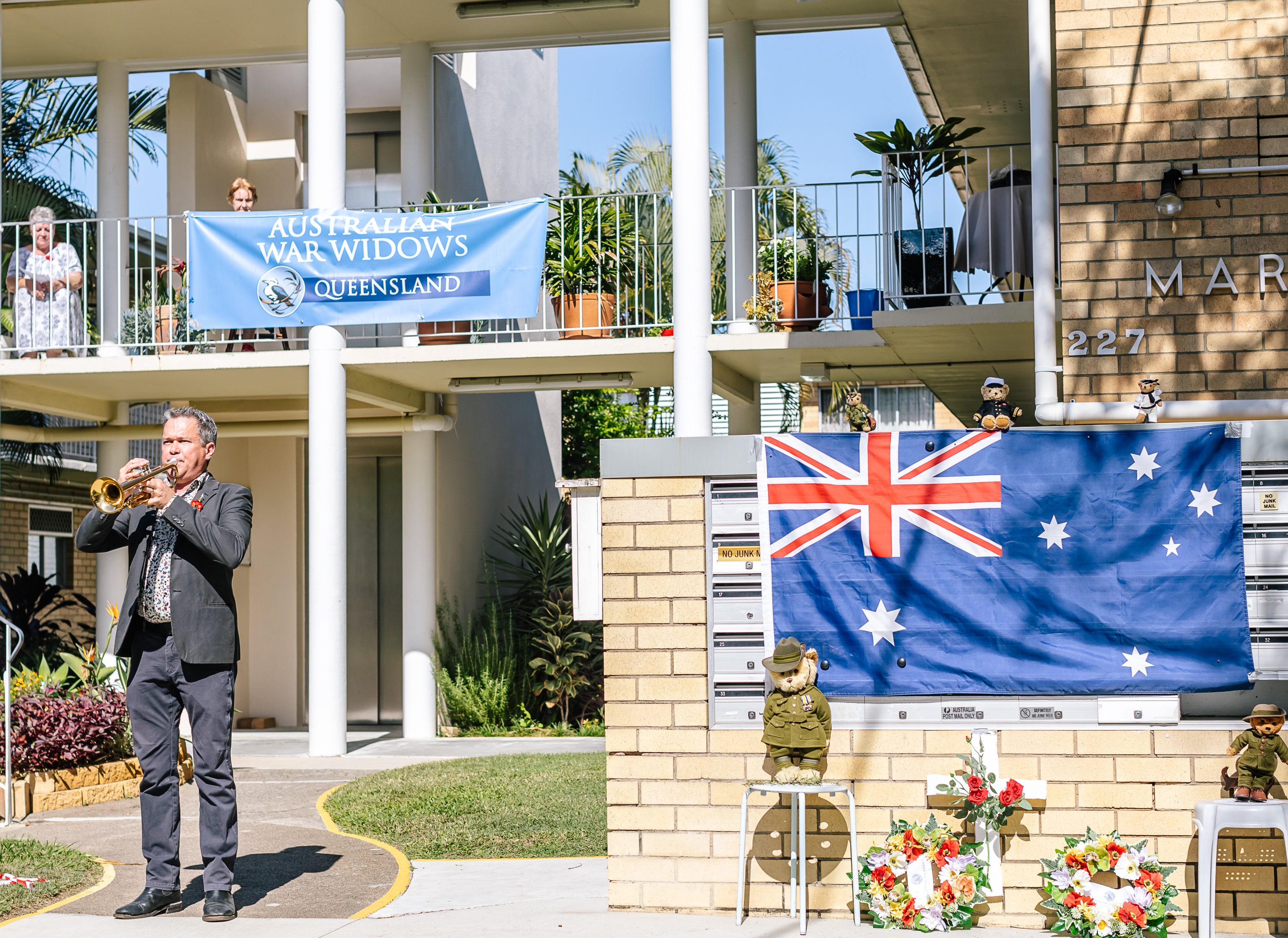 Alastair Tomkins, wearing a grey suit, plays the Last Post on a trumpet on Anzac Day 2020. He is playing to the war widows residents of a Marina Court unit complex in New Farm, Brisbane.