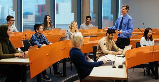 9 students of different ages and backgrounds sit at desks in a University of Newcastle room, listening to a lecturer.