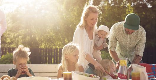 A family with a mother father and 3 young children are around an outdoor table having a picnic bbq style dinner as the sun is setting