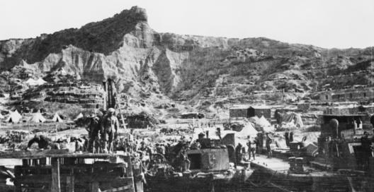 Men and boats crowd the docks with movement, at North Beach Gallipoli with the cliffs towering over them in the background 