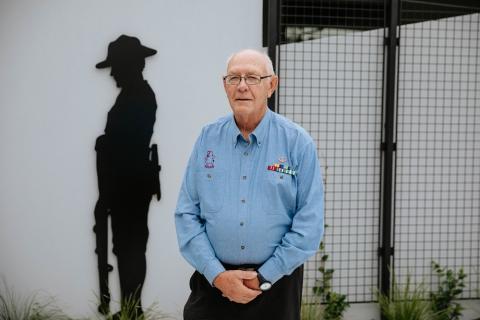 Elderly man wearing medal ribbons posing for camera standing next to Anzac sculpture